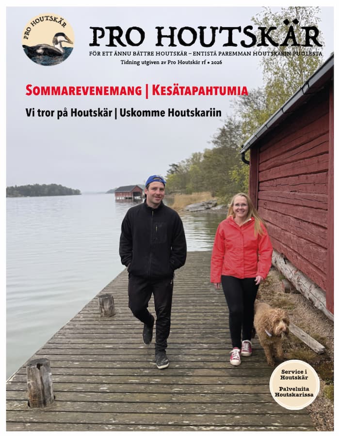 En man och en kvinna promenerar längs en träbrygga vid röda byggnader nära en lugn strand, vilket avbildas på omslaget till tidningen ”Pro Houtskär”.